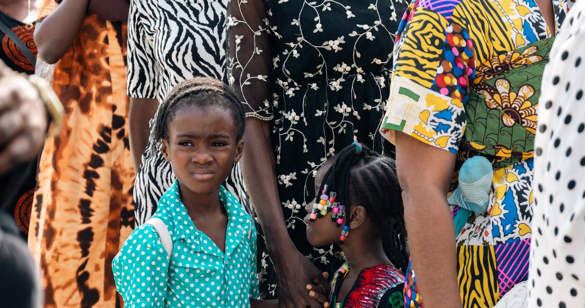 A girl in a teal patterned shirt looking towards the camera among adults waiting at the Ilasan outreach.