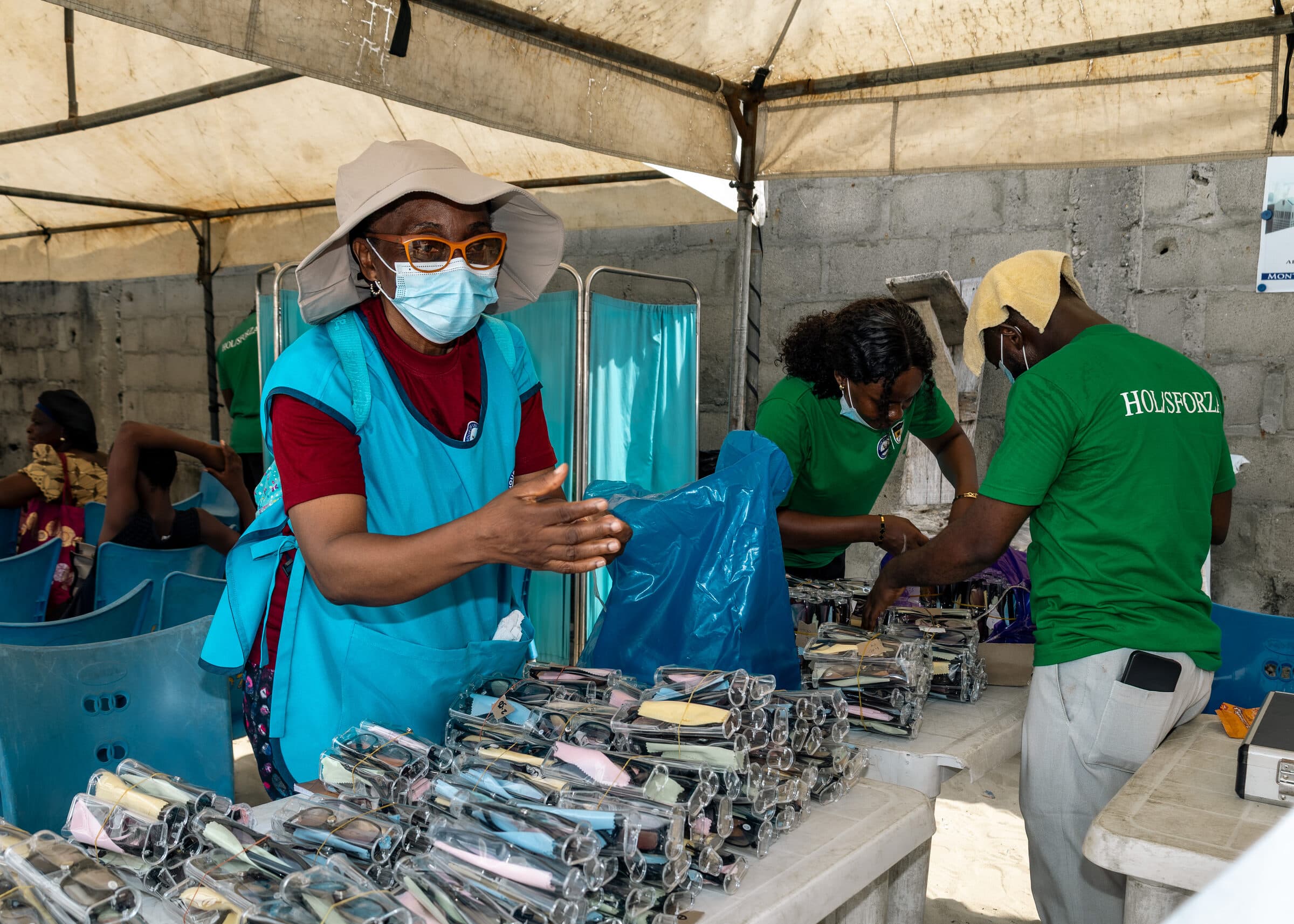 Volunteers in HOL and Sforza shirts sorting hundreds of packaged prescription eyeglasses on trestle tables under a canopy at the Ilasan medical outreach in Lekki, Lagos.