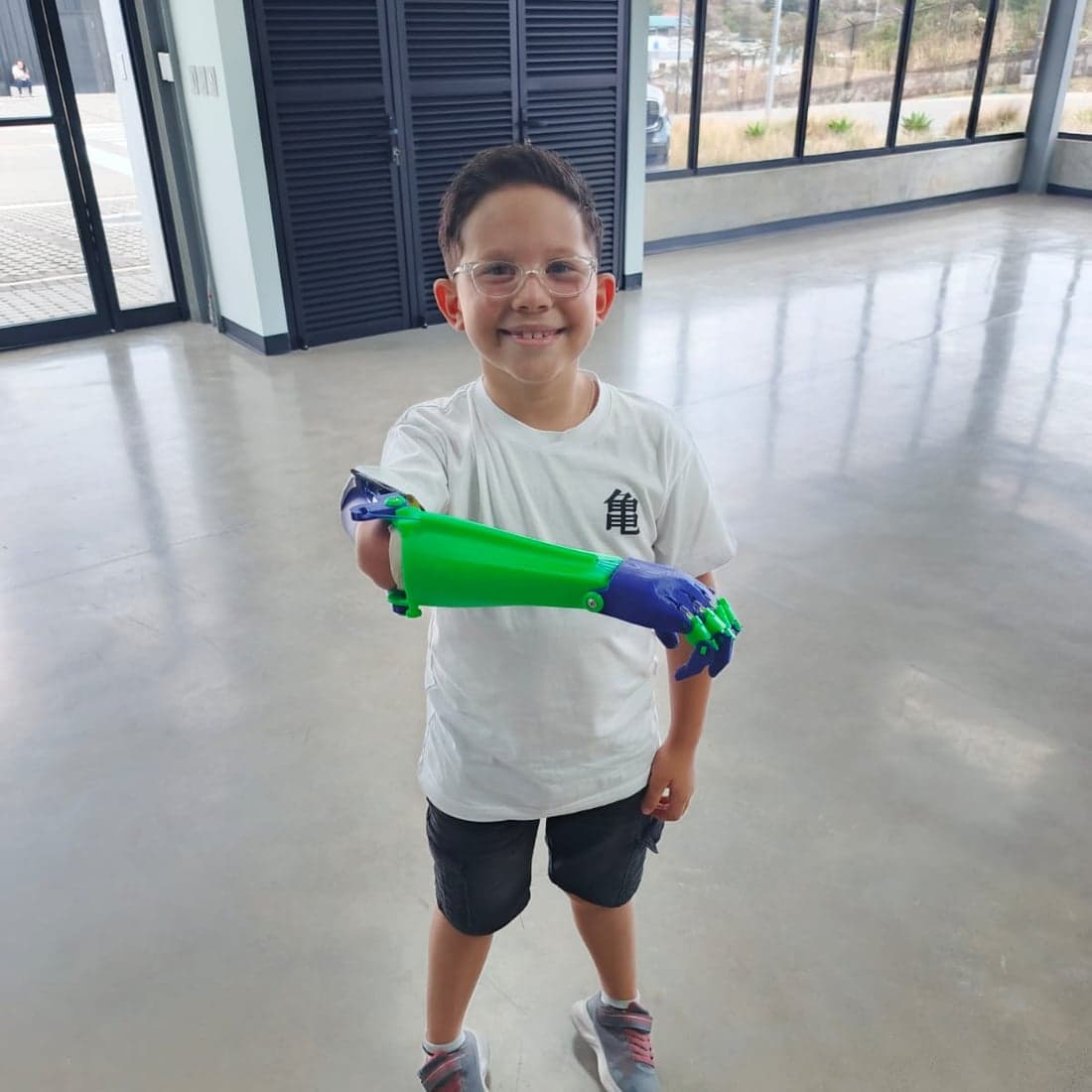 Nico standing and smiling, holding out his green and blue 3D printed forearm.